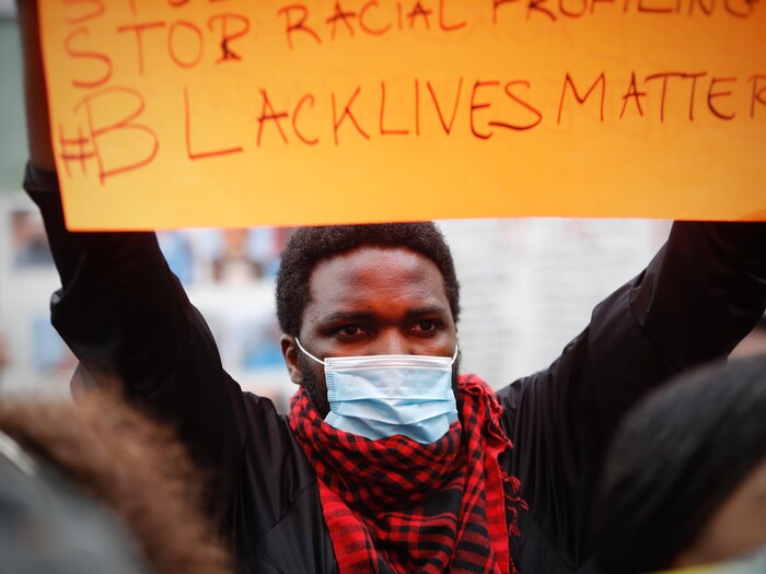 Un Noir avec un masque rouge tient une pancarte à deux mains. 