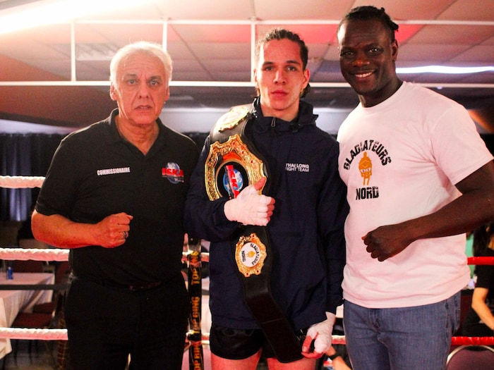 Le gagnant porte la ceinture de la victoire sur son épaule et est entouré du commissionnaire et de l'organisateur dans le ring.