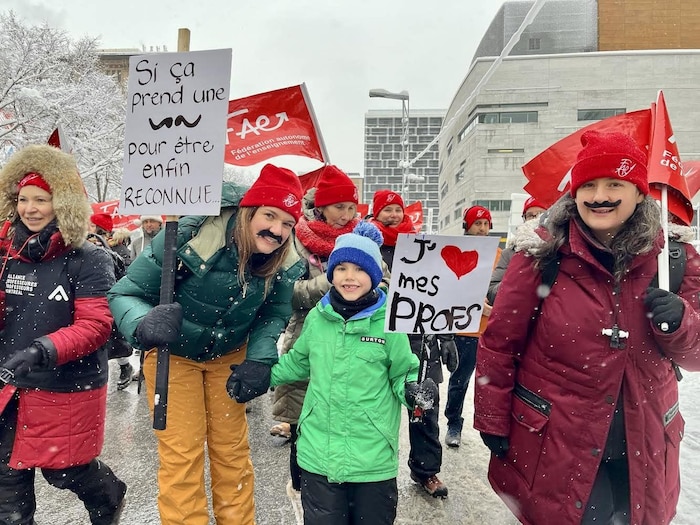 Une gréviste portant une moustache prend la pose en tenant la main d'un enfant. Leurs pancartes respectives indiquent : « Si ça prend une moustache pour être enfin reconnue... » et « J'aime mes profs ».
