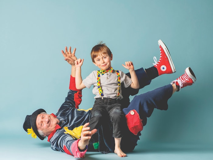 Un grand-père déguisé en clown est couché sur le dos, son petit-fils assis sur lui, tous les deux les bras dans les airs, souriants.