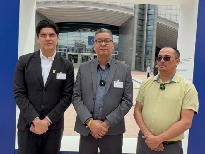 Trois hommes posent pour la caméra à l'extérieur, devant une grande bâtisse.