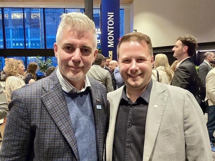Deux hommes posent en souriant dans une salle remplie de gens.