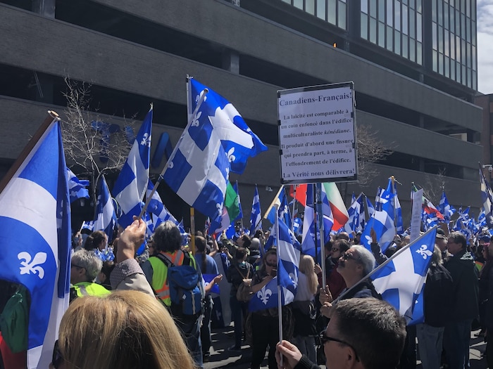 Des manifestants brandissent des drapeaux québécois et un autre brandit une pancarte s'adressant aux «Canadiens-Français».