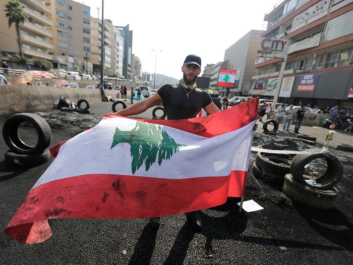 Un homme tient un drapeau libanais au milieu d'une rue où des pneus ont été incendiés. 