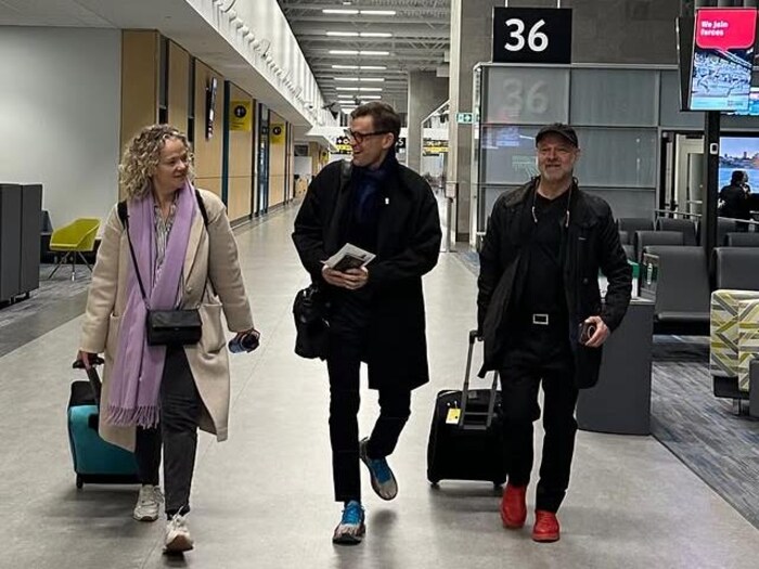 Les trois marchent avec leurs valises dans une allée de l'aéroport.