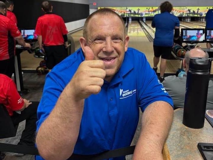 Un homme vêtu d'un polo bleu marqué « Équipe Québec » fait un signe de pouce levé au premier plan.
