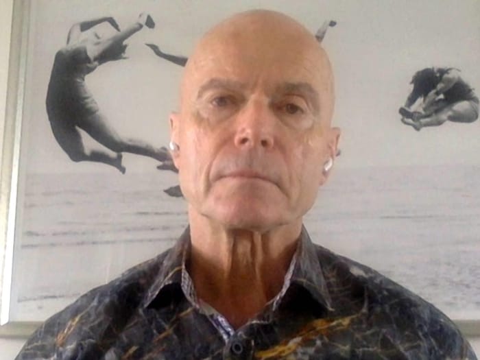 Un homme affiche un air neutre en regardant directement devant lui. Il y a derrière lui un cadre d'une photo en noir et blanc de gens qui plongent dans un lac.