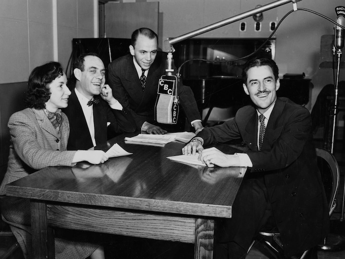 Dans un studio de radio, Lisette LeRoyer, Miville Couture, Roger De Vaudreuil et René Lecavalier, sont assis autour d'une table, un micro suspendu entre eux.