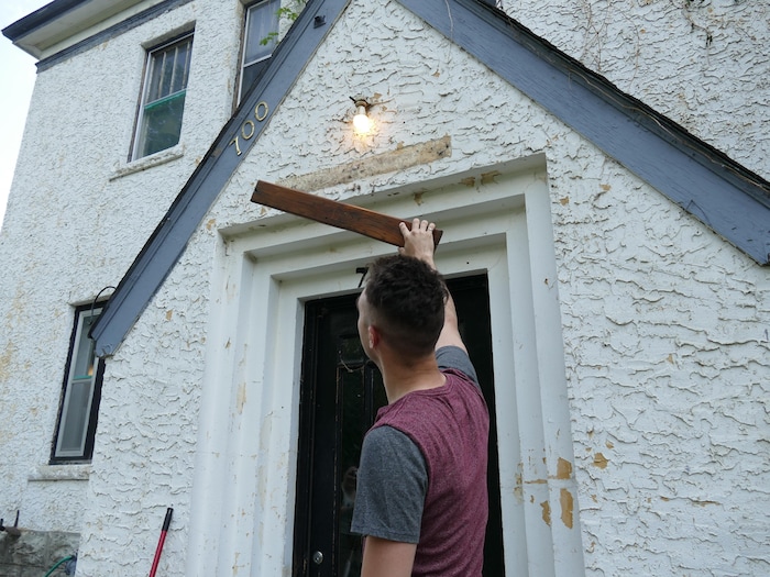 Un homme tient une petite planche de bois devant l'entrée d'une maison.