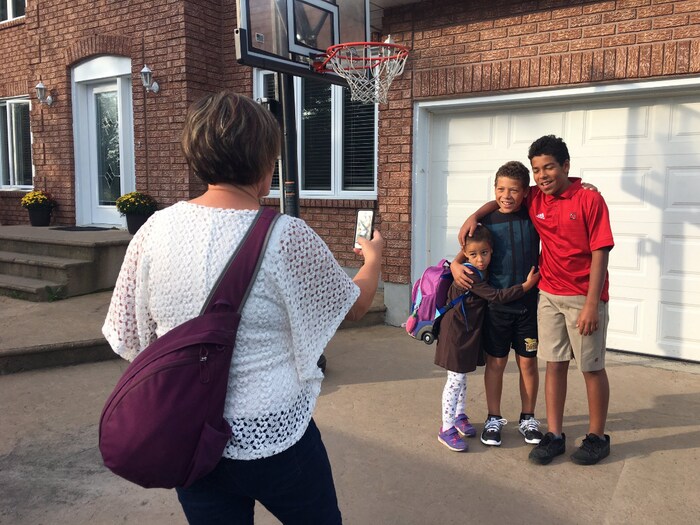 Une femme prend une photo de trois enfants devant une maison.