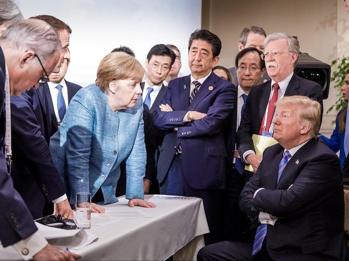Donald Trump, assis, semble se faire gronder par Angela Merkel, debout devant lui. 