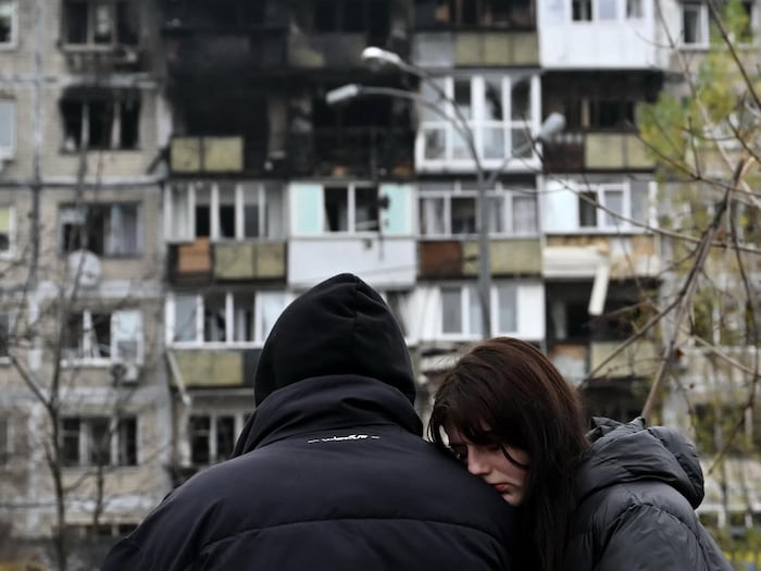 Une femme appuie son visage sur l'épaule de son compagnon devant un immeuble endommagé par une frappe.