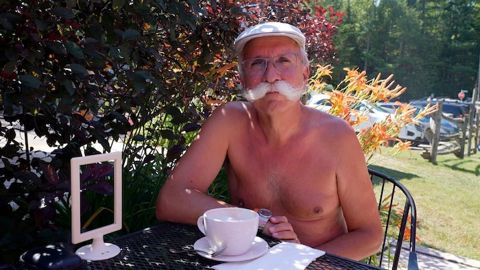 Le président de la Fédération de naturisme Stéphane Deschênes pose pour la camera, assis a une table devant son café. 
