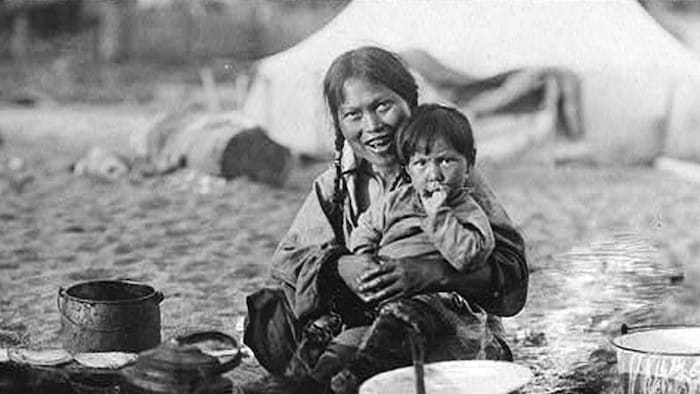 Photographie d’une femme tenant un enfant dans ses bras. Elle est entourée de chaudrons et d’ustencils de cuisine en plein air.