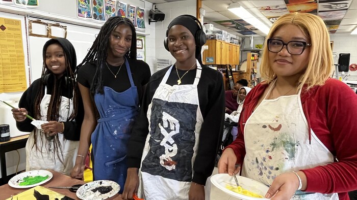 Quatre jeunes filles souriantes, en tablier, dans une salle de classe tiennent des pinceaux et des pochoirs de peinture.