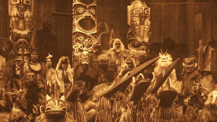 Photo d’un groupe de personnes en tenue d’apparat traditionnelle devant des mâts-totems.