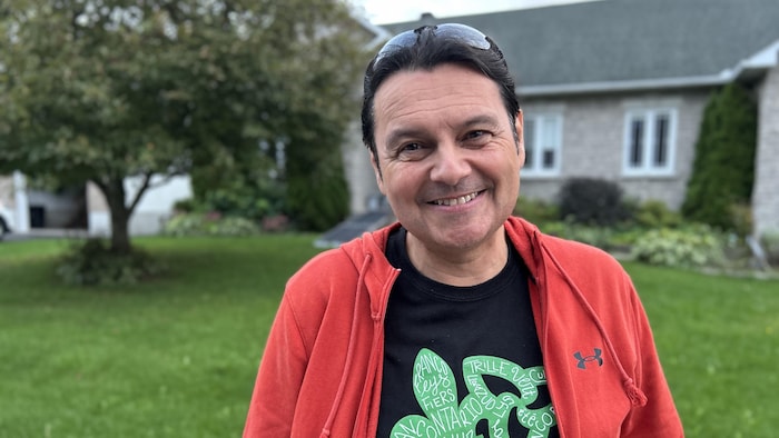 Un homme souriant aux cheveux long devant un arbre et une maison.