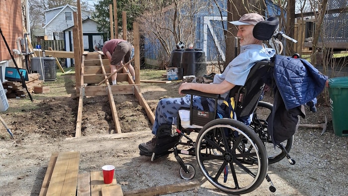 Un homme en fauteuil roulant observe les travaux qui sont faits sur sa maison.