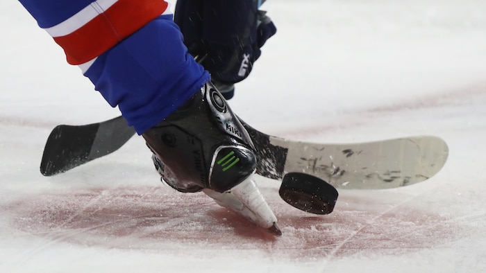 Deux bâtons de hockey se disputent la rondelle entre des patins.
