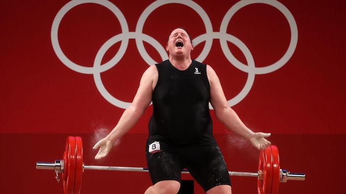 Une femme pousse un cri dans une épreuve d'haltérophilie. L'emblème des Jeux olympiques est en arrière-plan.