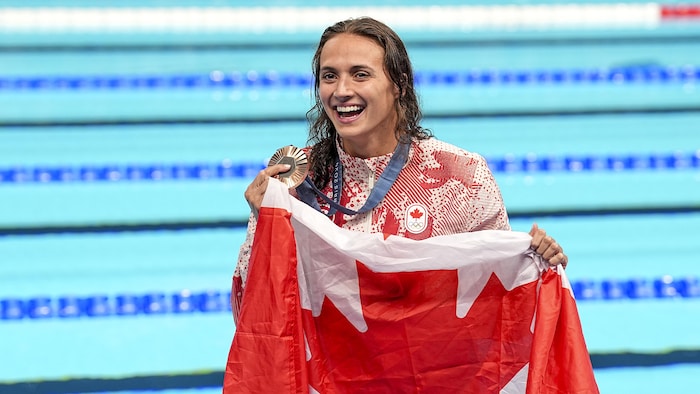 Une femme tient sa médaille de bronze et un drapeau du Canada en souriant devant la piscine.