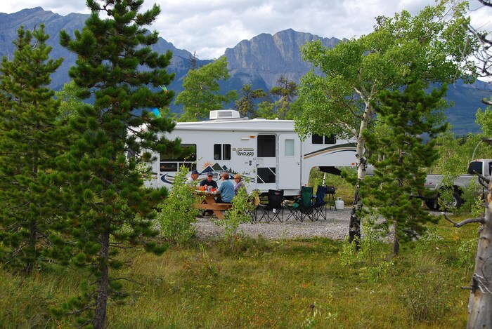 Quatre campeurs sont installés à une table de pique-nique, devant une caravane installée dans un terrain de camping de montagne.