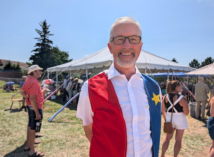 Debout en plein air devant un chapiteau, un homme porte une veste rouge et bleue par-dessus une chemise blanche, pour symboliser le drapeau de l'Acadie.
