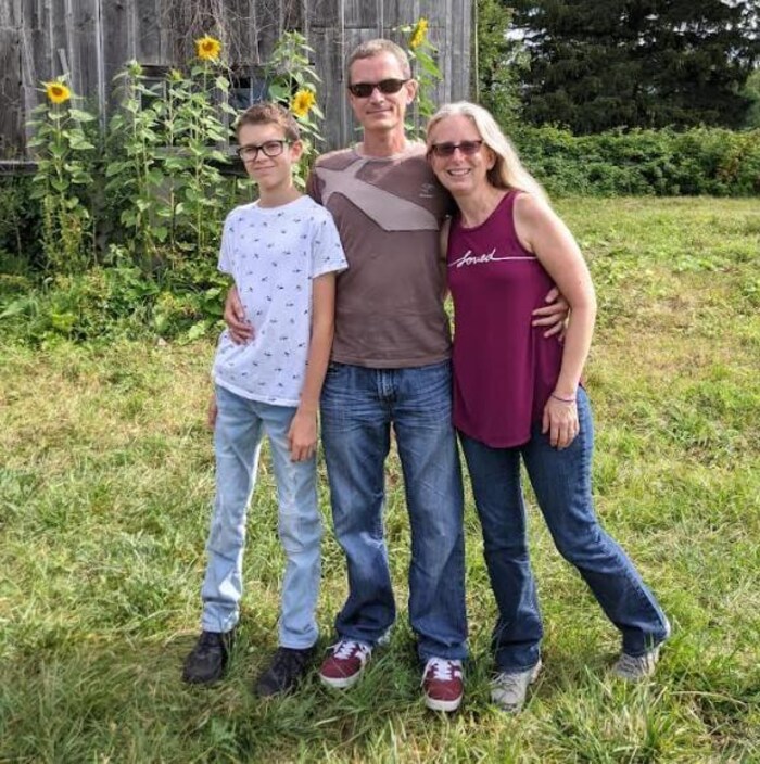 A mother, father and child standing on the lawn, by a wooden building.