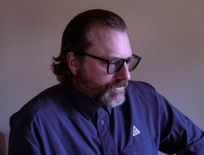 Un homme assis, tourné vers la droite, le regard dirigé vers le bas. Il porte des lunettes noires, une barbe et a les cheveux foncés lissés vers l'arrière.