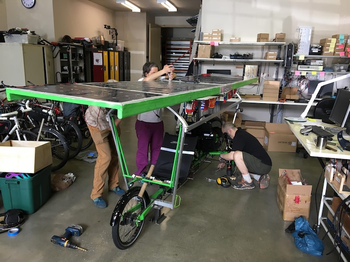 Trois personnes réparent un vélo avec un panneau à énergie solaire dans un atelier.