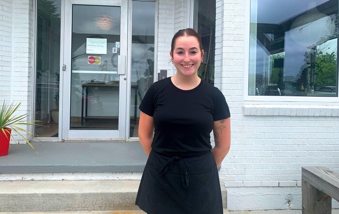 Une femme habillée en noir, debout les deux mains dans le dos, pose pour une photo en faisant un grand sourire, devant un bâtiment à la façade de briques peinte en blanc.