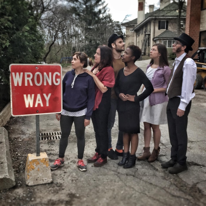 5 acteurs regardent dans une même direction alors qu'un autre regarde dans le sens opposé. Sur un panneau d'affichage est inscrit en anglais : Wrong Way, mauvaise direction en français.
