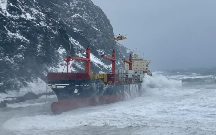 Un navire près de rochers, de grosses vagues, un hélicoptère au-dessus du bateau, dans une température hivernale inhospitalière.