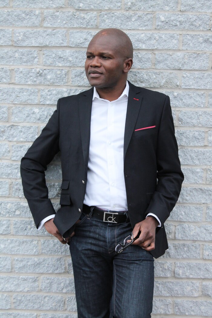 Portrait du juriste et écrivain d'origine congolaise Blaise Ndala, debout en veston et chemise blanche devant un mur de briques gris pâle. 