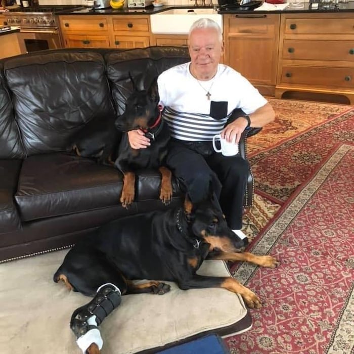 Un septuagénaire vêtu d'un chandail blanc, une petite croix autour du cou, est assis sur un divan en cuir à côté d'un chien doberman. Un autre doberman est couché par terre avec une patte dans le plâtre.