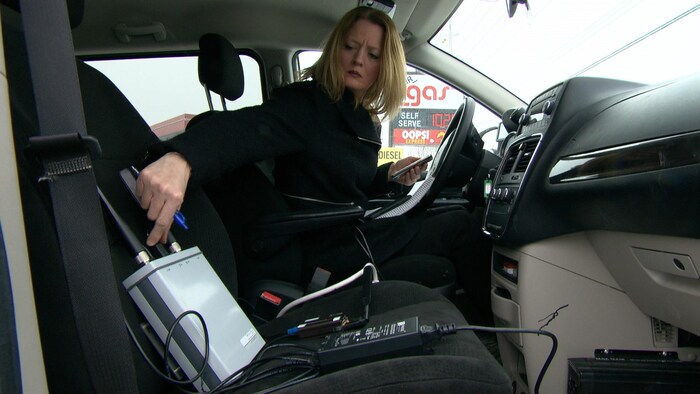 La journaliste Brigitte Bureau assise dans une voiture à côté d'un appareil pouvant déceler la présence d'intercepteurs d'IMSI.