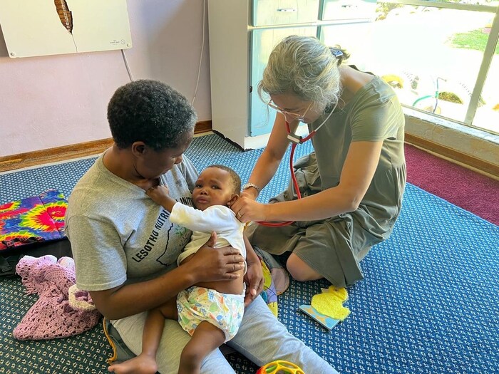 A woman puts a stethoscope to a baby's back. The mother of the child watches.