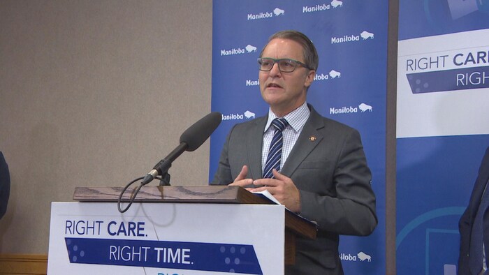 Un homme, avec des lunettes, debout devant un présentoir et un micro. En arrière, un panneau bleu du gouvernement du Manitoba qui dit « Right Care, Right Time ».