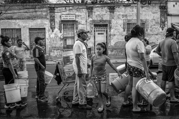 Des hommes, des femmes et des enfants attendent en file avec des seaux vides pour recueillir de l'eau, dans une rue du Mexique.