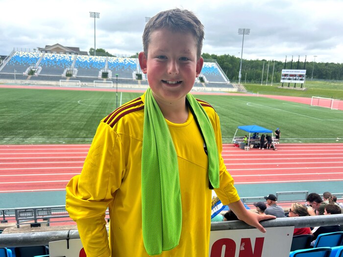 Un adolescent, vêtu d'un uniforme jaune de gardien de but, une serviette verte posée autour du cou, est debout le dos à un terrain de soccer et sourit pour une photo.