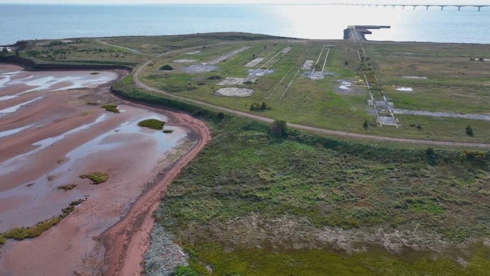 Photo aérienne d'un terrain vacant, couvert de végétation et de béton, près d'une rive couverte d'une terre rouge et boueuse. Le pont de la Confédération apparaît dans le lointain, en haut de l'image.