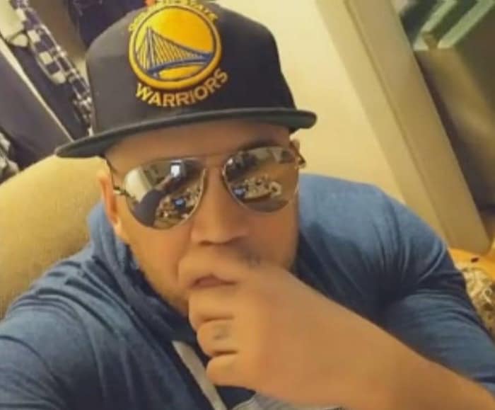 Un homme assis dans un fauteuil. Il porte une casquette des Golden State Warriors, des lunettes fumées et sa main gauche couvre partiellement sa bouche.