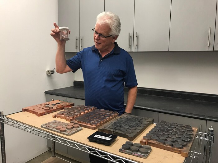 Un homme tient un bocal dans sa main droite. Devant lui, il y a une étagère sur laquelle reposent de petites plaquettes d'acier. Il s'agit de Christian Babin.