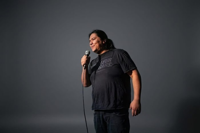 Microphone à la main, un humoriste sourit sous les projecteurs devant un fond sombre.