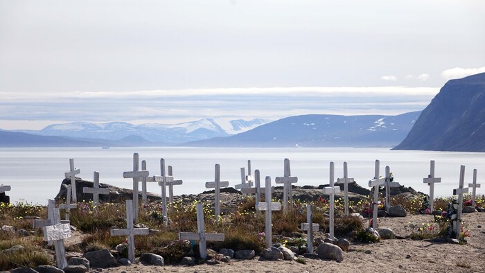 De nombreuses croix blanches sont posées sur le bord d'une plage. De loin, derrière les croix, on peut voir à travers le brouillard une immense étendue d'eau, des montagnes et de la neige. 
