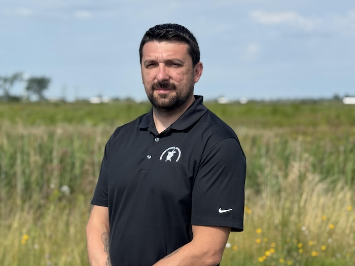 Un homme vêtu d’un polo noir se tient devant un vaste champ couvert d’herbes hautes et de fleurs jaunes.