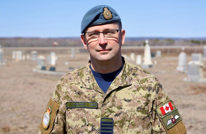 Un militaire en uniforme, portant un képi bleu et des lunettes, est debout en plein air. On distingue les tombes d'un cimetière, floues à plusieurs mètres derrière lui.