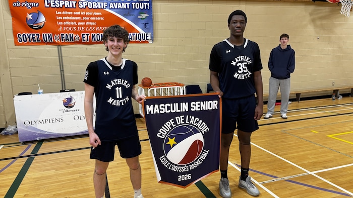 Two teenagers in blue and white uniforms pose proudly with a banner on the playing surface in a gymnasium.