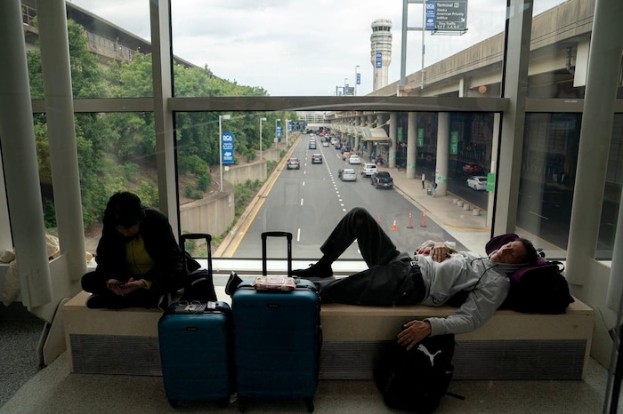 Two people, one sitting and the other, lying down, on a bench in an airport hall overlooking a road and trees.