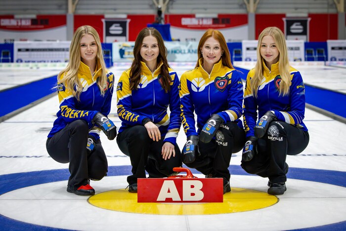 Photo de groupe des quatre membres de l'équipe féminine de curling de l'Alberta, en uniforme noir, bleu et jaune. Elles sont agenouillées sur la glace devant une pierre de curling.